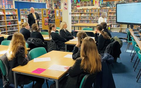 A group of students aged approximately 14, receive training from academy staff on being reading mentors. Image taken in the academy library with shelves of colourful books and reading tables.