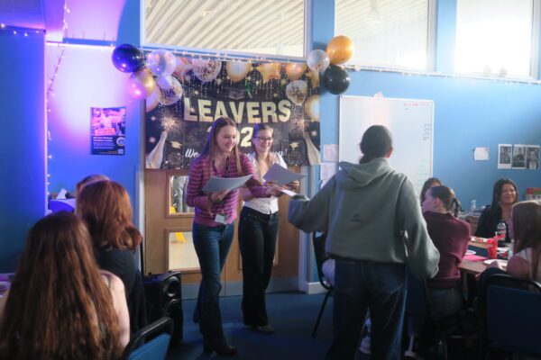 Two girls in front of a leavers banner at sixth form, receiving certificates.